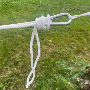 A close-up of a white braided rope tied with a knot and attached to a silver metal carabiner on one end and Europe Bound Hammock on the other, set against a blurred green grassy outdoor background. | ScoutTech Outfitters