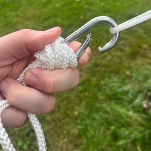 A close-up of a hand holding a white braided rope knot with a silver metal carabiner clipped onto it, outdoors with blurred green grass in the background.  The rope is an attachment point to suspend a Europe Bound 1 Person Hammock.  | ScoutTech Outfitters