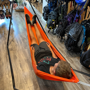 A person lying flat in a bright orange Europe Bound camping hammock suspended over a wood floor, with backpacks hanging on the wall behind them in a retail store setting. | ScoutTech Outfitters