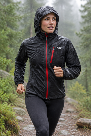 A woman in her 20's is shown running in the woods along a trail in the rain.  She expresses enjoyment despite the weather as she is wearing her Outdoor Research Women's Helium II Ultralight Full Zip Rain Jacket in Nocturnal Black with Red Zippers and a reflective OR logo on the left chest above a zippered pocket. | ScoutTech Outfitters